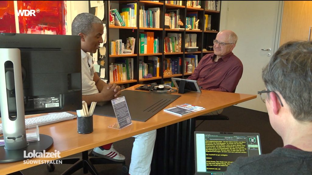 Das Sprechzimmer eines Arztes. Arzt und Patient sitzen sich gegenüber. Vor dem Patient setz ein Tablet auf dem Tisch. Die Schriftdolmetscherin sitzt ein Stück abseits. Man sieht ihren Hinterkopf und das Display ihres Laptops.