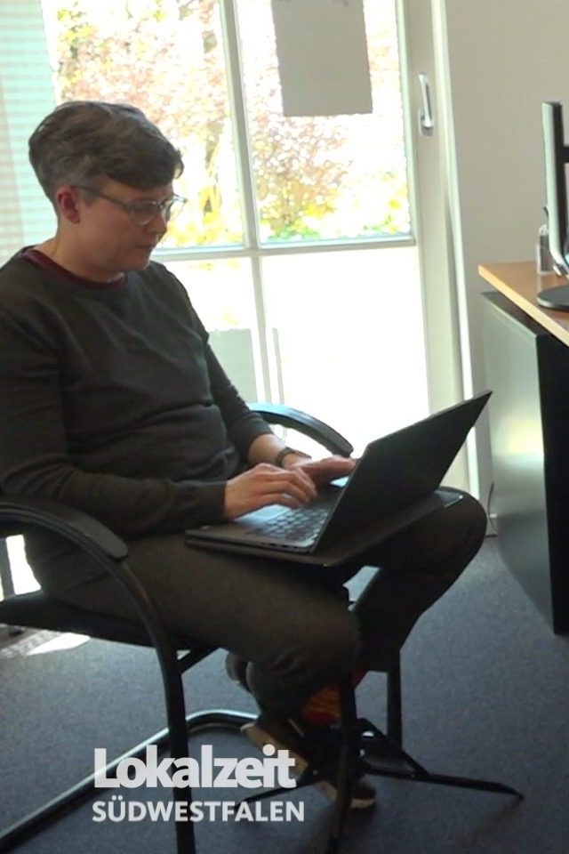 Die Schriftdolmetscherin sitzt auf einem Stuhl. Vor ihr steht ein kleines Stativ, darauf liegt ein aufgeklapptes Laptop, auf dem sie tippt.