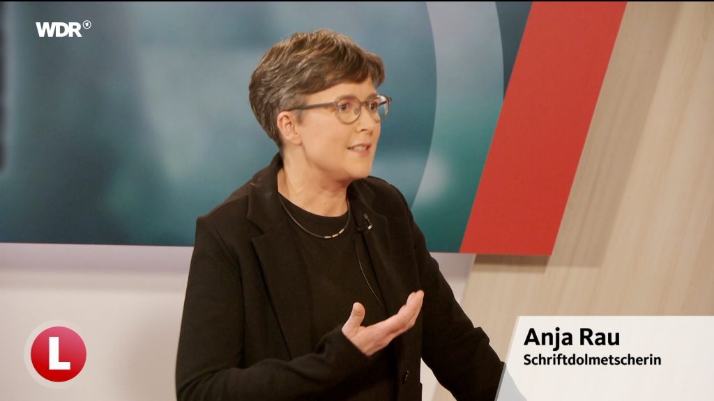Anja Rau, Schriftdolmetscherin aus Siegen in der WDR Lokalzeit Südwestfalen. Eine Frau vor einem Studio-Hintergrund, kurze braune Haare, Brille, schwazes-T-Shirt und schwarzes Jackett.